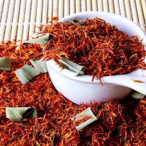 May include: A white ceramic scoop overflowing with vibrant red-orange saffron threads and green leaves. The saffron is scattered across a bamboo mat, creating a visually appealing display of culinary ingredients.