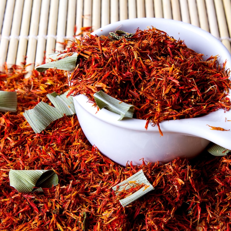 May include: A white ceramic scoop overflowing with vibrant red-orange saffron threads and green leaves. The saffron is scattered across a bamboo mat, creating a visually appealing display of culinary ingredients.