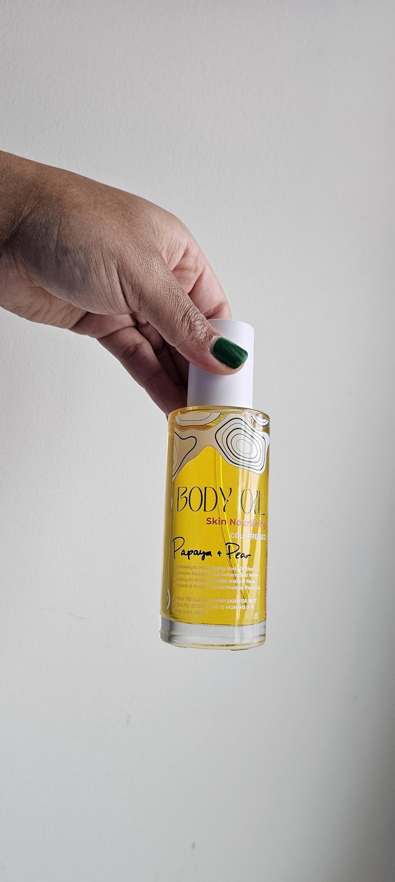 May include: A clear glass bottle of body oil with a white cap, held by a hand with green nail polish. The label reads "BODY OIL" and "Papaya + Pear". The oil appears yellow.