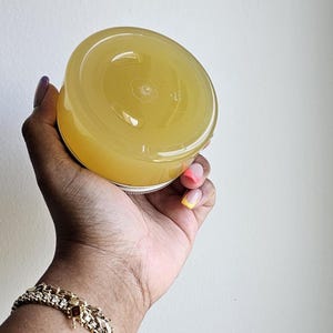 May include: A clear jar filled with a yellow liquid, held in a hand with a gold bracelet. The jar has a silver lid and is viewed from above. The hand has a colorful manicure with pink, orange, and yellow nail polish.
