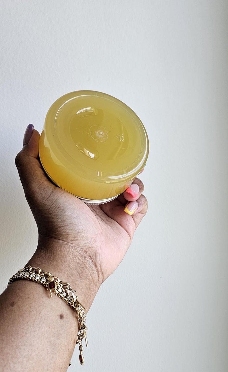 May include: A clear jar filled with a yellow liquid, held in a hand with a gold bracelet. The jar has a silver lid and is viewed from above. The hand has a colorful manicure with pink, orange, and yellow nail polish.