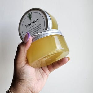 May include: Two clear plastic jars filled with a yellow liquid, held in a hand. The jars have silver lids and labels that read "Rosemary." The text on the label also mentions use as a pre-shampoo or overnight hair mask. A gold bracelet is visible on the wrist.