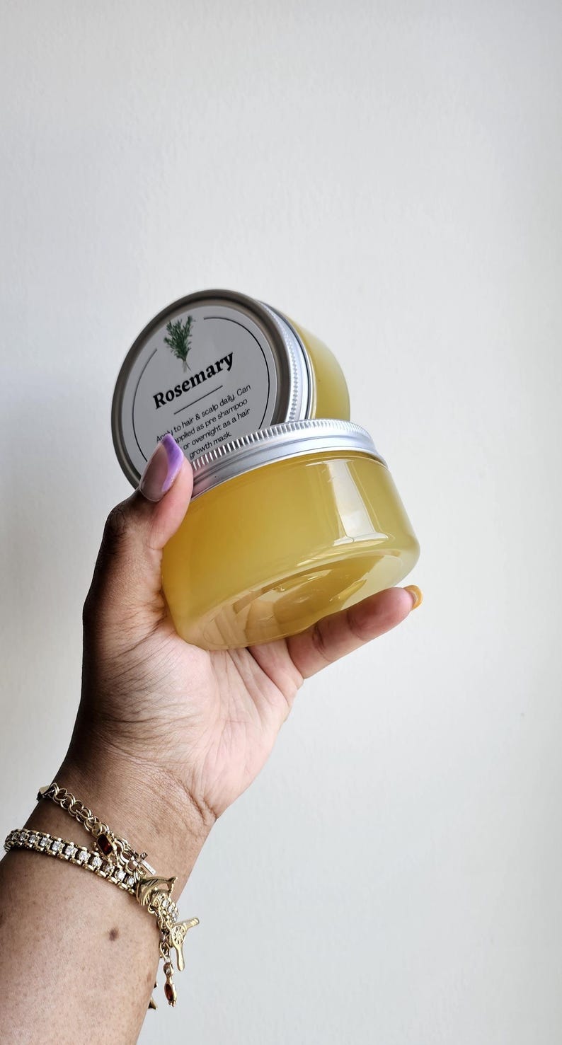May include: Two clear plastic jars filled with a yellow liquid, held in a hand. The jars have silver lids and labels that read "Rosemary." The text on the label also mentions use as a pre-shampoo or overnight hair mask. A gold bracelet is visible on the wrist.