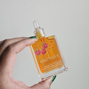 Puede incluir: Una botella de vidrio transparente de aceite de frambuesa prensado en fr&iacute;o. La botella tiene un cuentagotas y una etiqueta con la palabra "RASPBERRY" y un dibujo de frambuesas. El aceite es de color dorado.