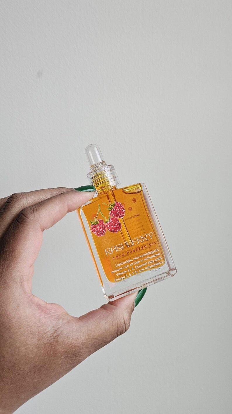 Puede incluir: Una botella de vidrio transparente de aceite de frambuesa prensado en fr&iacute;o. La botella tiene un cuentagotas y una etiqueta con la palabra "RASPBERRY" y un dibujo de frambuesas. El aceite es de color dorado.