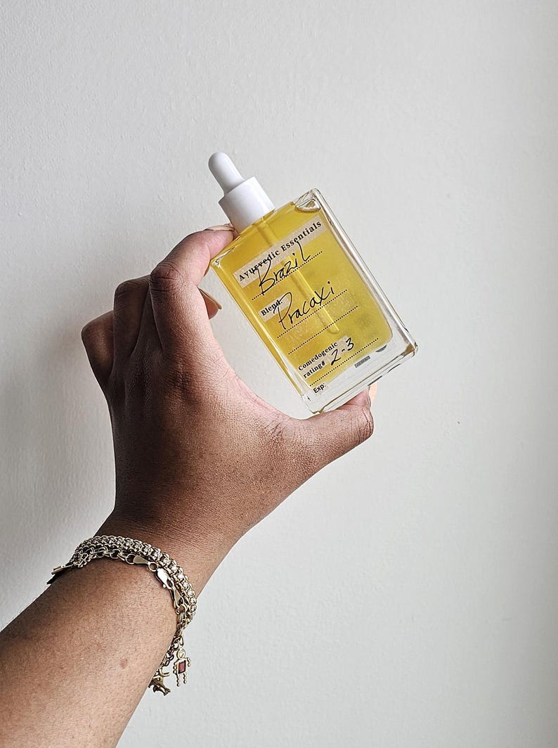 May include: A clear glass bottle with a dropper top filled with a yellow liquid. The bottle has a label that reads "Ayurvedic Essentials Brazil Pracaxi Blend Comedogenic rating 2-3 Exp."