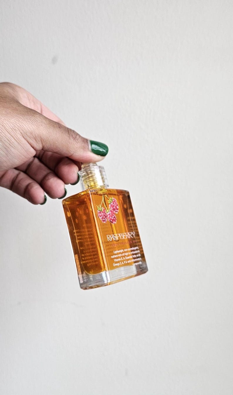 Puede incluir: Una botella de vidrio transparente llena de l&iacute;quido &aacute;mbar, etiquetada como "Raspberry". La etiqueta presenta una ilustraci&oacute;n de frambuesa. La botella se sostiene sobre un fondo blanco.