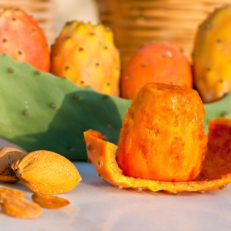 May include: Close-up of prickly pear fruit, peeled to reveal the bright orange flesh. Several whole fruits and a green cactus pad are in the background. Almonds are in the foreground. The fruits are orange and yellow.