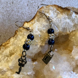 May include: A pair of dangle earrings with a key and a padlock charm. The earrings feature black beads with gold accents.