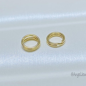 Op de afbeelding: Twee goudkleurige dubbele sprongringen, waarschijnlijk voor het maken van sieraden, worden getoond tegen een witte, licht getextureerde achtergrond. De ringen zijn cirkelvormig met een opening voor bevestiging. De afbeelding is goed verlicht.