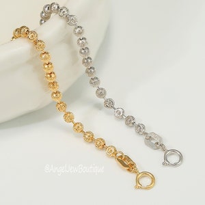 Op de afbeelding: Twee kralenarmbanden, een gouden en een zilveren, met een sluiting. De armbanden liggen op een wit oppervlak.