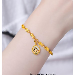 May include: A gold-toned chain bracelet featuring a series of small, repeating decorative links and a single oval pendant. The pendant displays a colorful enamel illustration of a character with dark hair and a blue outfit set within a gold frame.