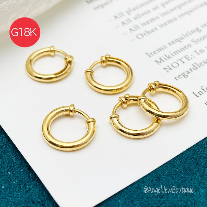 May include: Four gold-toned hoop earrings with a polished finish. The earrings are circular with a hinged closure mechanism. A red circular graphic in the top left corner contains the white text G18K. The jewelry rests on a white paper surface.