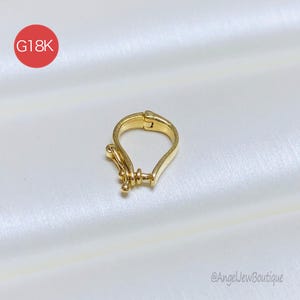 May include: A gold-coloured jewellery clasp with a distinctive design. The clasp is teardrop-shaped with a hinged closure. The image includes a red circle with the text "G18K". The background is a white, silky fabric.