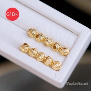 May include: Close-up of several gold-colored, textured beads arranged in rows within a white tray. The beads have a detailed, ridged design. A red circle with the text "G18K" is in the upper left corner.
