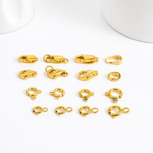 May include: A collection of gold-colored jewelry clasps and connectors, including lobster claw clasps, spring ring clasps, and other styles. Some clasps feature small blue gemstones. The clasps are arranged on a white surface.