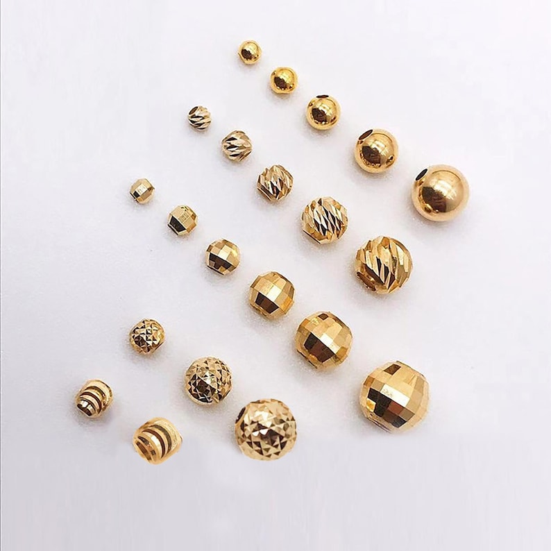 Peut inclure: Une collection de perles dor&eacute;es de diff&eacute;rentes tailles et formes, dont des mod&egrave;les &agrave; facettes, ronds et cylindriques. Les perles sont dispers&eacute;es sur une surface blanche, mettant en valeur leurs surfaces r&eacute;fl&eacute;chissantes.