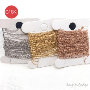 May include: Three spools of delicate chain in silver, gold, and rose gold. Each chain is neatly wound on a white card. The upper left corner has a red circle with the text "G18K". The image is against a white background.