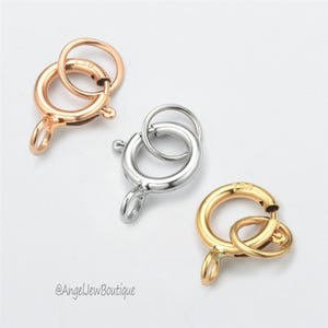 May include: Three metal spring ring clasps in rose gold, silver, and gold. Each clasp has a circular ring and a small loop for attaching jewelry. The clasps are on a white background. The brand name "@AngelJewBoutique" is in the bottom left corner.