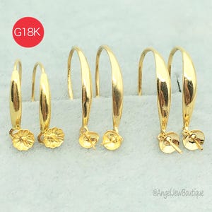 Könnte beinhalten: Mehrere Paare goldfarbener Creolen mit floralem Design am unteren Ende. Die Ohrringe haben eine gebogene, längliche Form. Ein roter Kreis mit dem Text "G18K" befindet sich in der oberen linken Ecke. Die Ohrringe werden auf einer hellgrauen Oberfläche präsentiert.