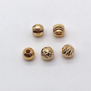 Peut inclure: Une collection de cinq perles dor&eacute;es. Les perles pr&eacute;sentent diff&eacute;rentes formes, notamment des facettes, des formes rondes et cylindriques. Les surfaces r&eacute;fl&eacute;chissent la lumi&egrave;re, mettant en valeur leur &eacute;clat m&eacute;tallique. Ces perles sont adapt&eacute;es &agrave; la cr&eacute;ation de bijoux.
