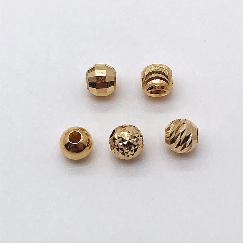 Peut inclure: Une collection de cinq perles dor&eacute;es. Les perles pr&eacute;sentent diff&eacute;rentes formes, notamment des facettes, des formes rondes et cylindriques. Les surfaces r&eacute;fl&eacute;chissent la lumi&egrave;re, mettant en valeur leur &eacute;clat m&eacute;tallique. Ces perles sont adapt&eacute;es &agrave; la cr&eacute;ation de bijoux.