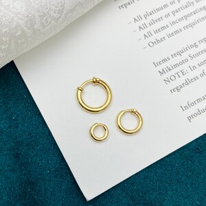 May include: Three gold hoop earrings of varying sizes arranged in a cluster on a white paper surface. Each earring features a simple circular band design with a small hinge mechanism, showcasing a polished metallic finish and a consistent yellow gold tone.