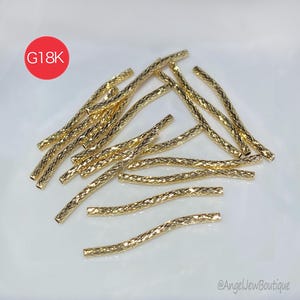 Puede incluir: Una colección de tubos metálicos dorados y texturizados dispersos sobre una superficie blanca. Los tubos tienen un patrón acanalado y una ligera curva. Un círculo rojo con el texto "G18K" está en la esquina superior izquierda. El nombre de la tienda, "@AngelNewBoutique", está en la esquina inferior derecha.