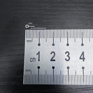 May include: A small, silver-colored metal pin with a circular head, measuring 6.5mm in length and 0.8mm in thickness. The pin is placed next to a ruler for scale, with markings in centimeters and millimeters. The background is a dark, textured surface.