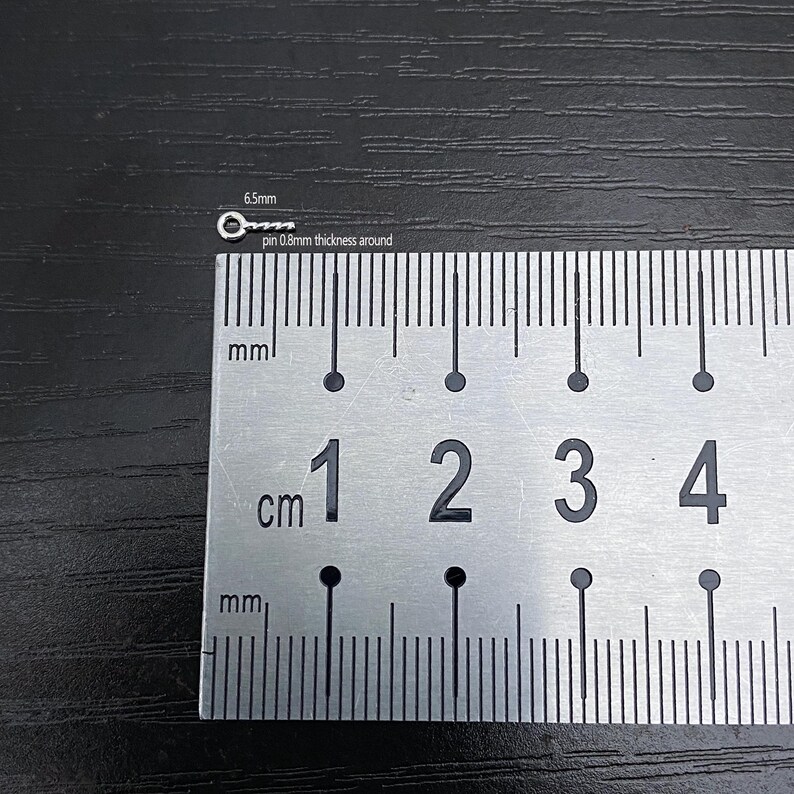 May include: A small, silver-colored metal pin with a circular head, measuring 6.5mm in length and 0.8mm in thickness. The pin is placed next to a ruler for scale, with markings in centimeters and millimeters. The background is a dark, textured surface.