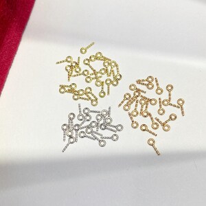 May include: A collection of small eye pins in gold, silver, and bronze tones. Each pin features a looped eye and a textured, straight post. The pins are scattered on a white surface, with a hint of red fabric in the corner.
