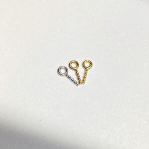 May include: Three metal eye pins in silver, gold, and gold-tone. Each pin has a looped eye and a twisted or braided stem. The pins are arranged on a white surface.