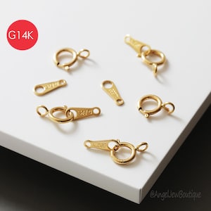 May include: Gold-colored jewelry clasps and connectors are arranged on a white surface. The clasps include spring ring clasps and lobster claw clasps. A red circle with the text "G14K" is in the upper left corner.