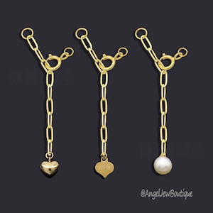 Può includere: Tre collane a catena color oro con diversi ciondoli. Una ha un ciondolo a forma di cuore, un'altra ha un ciondolo a forma di cuore con "Au750" inciso e la terza ha un ciondolo a perla bianca. Le catene sono su uno sfondo scuro.
