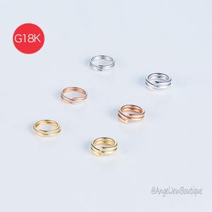 May include: A collection of small, delicate rings in gold, rose gold, and silver tones. The rings are arranged on a white surface. A red circle with the text "G18K" is in the upper left corner. The rings appear to be jewelry.