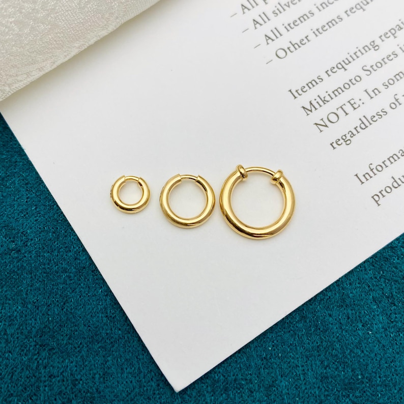 May include: Three gold-toned hoop earrings of varying sizes arranged in a horizontal row. Each earring features a smooth, circular band with a hinged closure mechanism, displaying a polished metallic finish and a simple, continuous design.