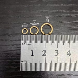 May include: Three gold-toned circular rings of varying sizes are displayed above a metal ruler. The rings are labeled small at 5.5mm, medium at 8mm, and big at 10.5mm. The ruler shows measurements in millimeters, providing a clear scale for the jewelry components.