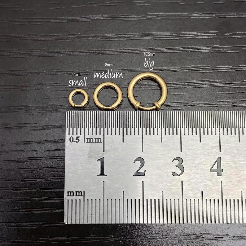 May include: Three gold-toned circular rings of varying sizes are displayed above a metal ruler. The rings are labeled small at 5.5mm, medium at 8mm, and big at 10.5mm. The ruler shows measurements in millimeters, providing a clear scale for the jewelry components.