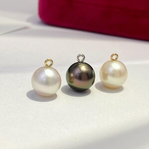 May include: Three lustrous pearl pendants. The pearls vary in color, with one white, one dark iridescent, and one cream-colored. Each pearl has a small metal loop at the top for hanging. The background is a soft white surface with a hint of a red box.