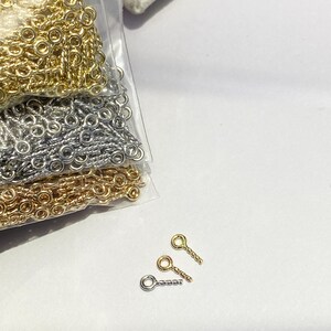 May include: Close-up of jewelry-making supplies. The image shows three small, gold-colored, silver-colored, and bronze-colored eye pins. Several bags of gold, silver, and bronze-colored chain are also visible.