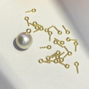 May include: A collection of small, gold-colored jewelry findings, including eye pins and a large, lustrous white pearl. The eye pins have a looped end and a textured stem. The pearl has a smooth, round shape and a subtle sheen.