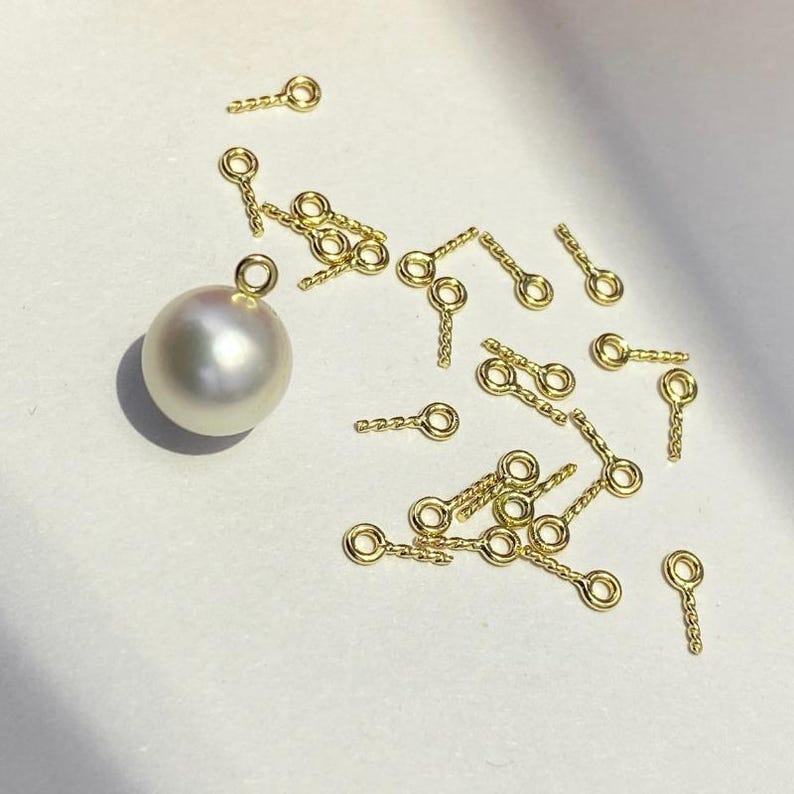 May include: A collection of small, gold-colored jewelry findings, including eye pins and a large, lustrous white pearl. The eye pins have a looped end and a textured stem. The pearl has a smooth, round shape and a subtle sheen.