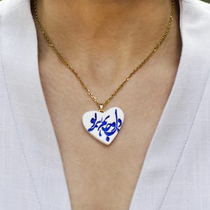 Könnte beinhalten: Eine goldene Kettenkette mit einem weißen herzförmigen Anhänger mit blauer arabischer Kalligraphie.