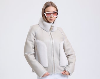 May include: A woman wearing a light gray shearling jacket with a white fur collar and white pants. She is wearing white sneakers and white sunglasses with pink lenses.