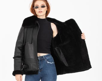 Könnte beinhalten: Schwarze Lederjacke mit Shearling-Futter und breitem Kragen. Die Jacke hat eine Schnallen-Detail und wird mit einem schwarzen Oberteil, blauen Jeans und schwarzen Absätzen kombiniert. Das Model trägt eine Sonnenbrille.