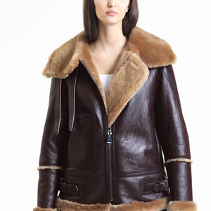 May include: A brown leather jacket with a shearling collar and trim. The jacket has a zip-up front, side pockets, and buckled cuffs. The model is wearing blue jeans and a white top.