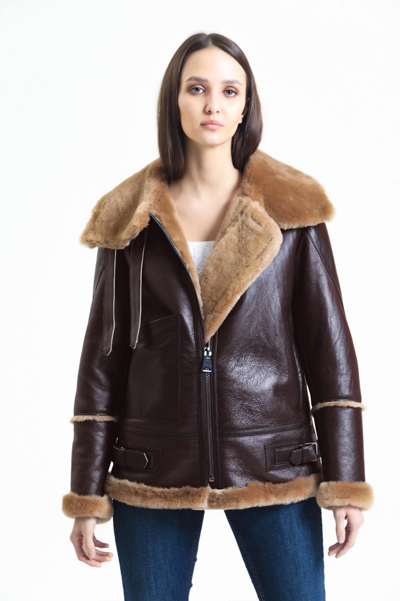 May include: A brown leather jacket with a shearling collar and trim. The jacket has a zip-up front, side pockets, and buckled cuffs. The model is wearing blue jeans and a white top.