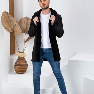 Puede incluir: Una chaqueta de piel de oveja negra con capucha. La chaqueta la lleva puesta una persona de pie sobre una plataforma blanca. La persona lleva pantalones vaqueros azules y zapatillas blancas.