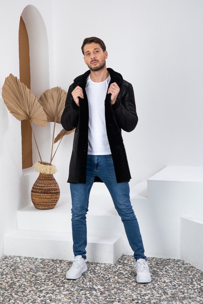 Puede incluir: Una chaqueta de piel de oveja negra con capucha. La chaqueta la lleva puesta una persona de pie sobre una plataforma blanca. La persona lleva pantalones vaqueros azules y zapatillas blancas.