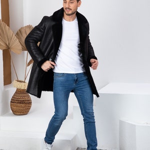 Puede incluir: Un hombre con un abrigo de piel de oveja negro, una camiseta blanca y pantalones vaqueros azules. Est&aacute; de pie en una plataforma blanca con un fondo blanco.